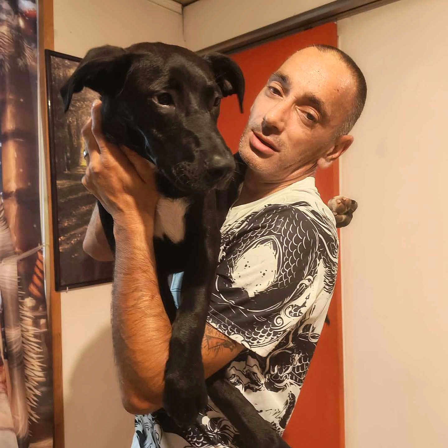 Princesse participe au concours pour gagner de l'argent avec cette photo : man, dog, indoor, holding, black_dog, short_hair, tattoo, tshirt, wall_art, door, portrait, pet, animal, casual, human, expression, arm, furniture, home, room