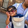 adult, baby_carrier, blue_shirt, brick_building, carrier, child, daylight, family, fashion, outdoor, parent, pink_clothing, portrait, smiling, street, sunglasses, toddler, travel, urban, window
