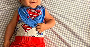 Kevin is registered to the contest to win money with this photo: baby, child, superman, superhero, smiling, lying_down, bed, bedspread, checkered_pattern, portrait, face, clothing, bib, red_shorts, blue_shirt, costume, indoors, happy, infant, photography