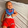 baby, child, superman, superhero, smiling, lying_down, bed, bedspread, checkered_pattern, portrait, face, clothing, bib, red_shorts, blue_shirt, costume, indoors, happy, infant, photography