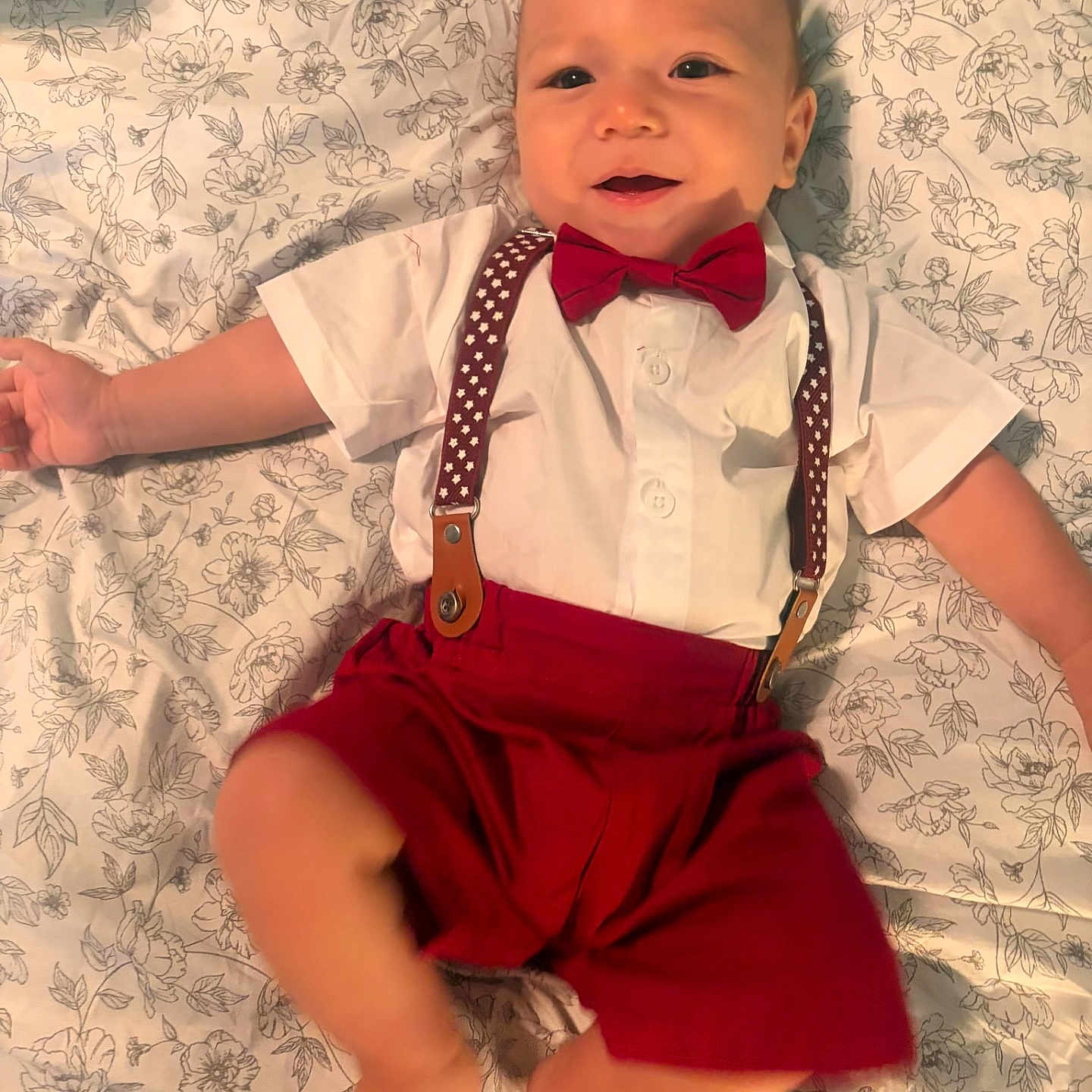 Kevin is registered to the contest to win money with this photo: baby, child, infant, smiling, portrait, lying_down, floral_blanket, bow_tie, suspenders, red_shorts, white_shirt, formal_outfit, happy, indoor, close_up, face, arms, legs, fashion, dapper