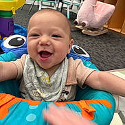 Kevin is registered to the contest to win money with this photo: baby, child, infant, smiling, happy, portrait, indoor, playroom, activity_seat, toy, rocking_horse, stuffed_animal, colorful, seated, arms_outstretched, clothing, bib, carpet, furniture, photography