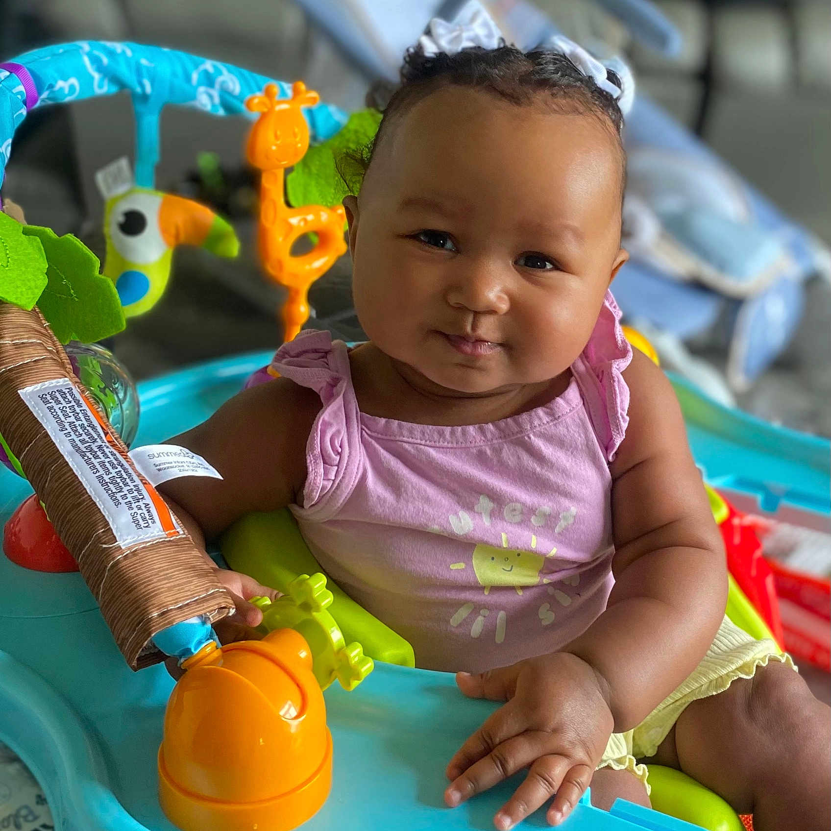 Noelle is registered to the contest to win money with this photo: activity_center, baby, baby_toys, child, chubby_cheeks, colorful, curly_hair, cute, face, happy, indoor, infant, person, pink_clothing, playtime, portrait, ribbons, seat, smile, yellow_clothing