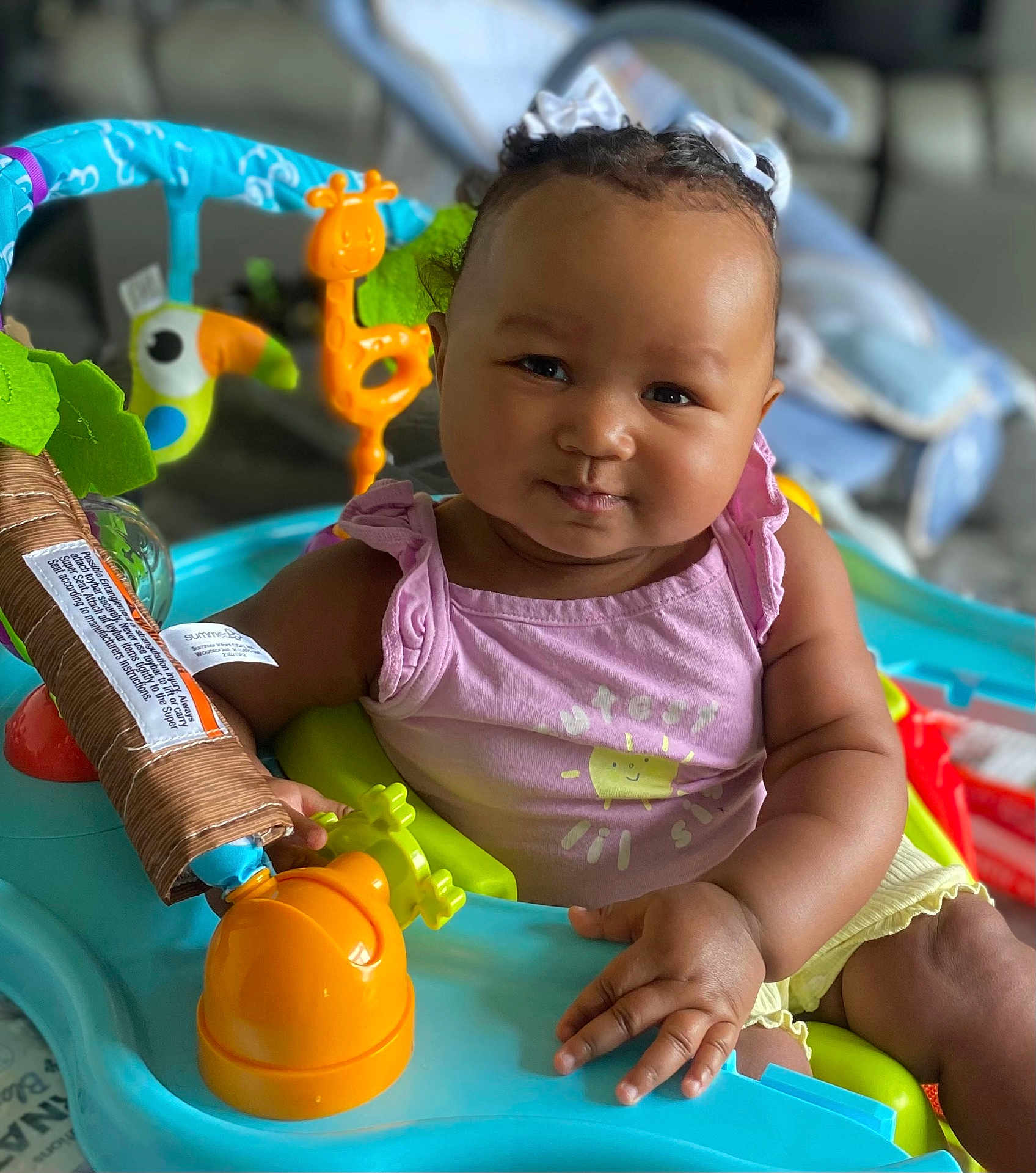 Noelle is registered to the contest to win money with this photo: baby, child, infant, face, smile, pink_clothing, yellow_clothing, baby_toys, activity_center, indoor, cute, playtime, ribbons, curly_hair, person, chubby_cheeks, seat, colorful, happy, portrait