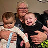 babies, grandmother, family, infants, smiling, portrait, indoors, couch, sitting, holding, caregiver, glasses, blonde_hair, black_clothing, floral_clothing, baby_bottle_absent, living_room, wall_clock, happy, family_photo