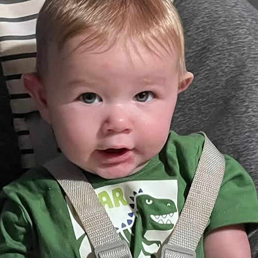 Emmett is registered to the contest to win money with this photo: baby, child, high_chair, sitting, portrait, face, wide_eyes, green_shirt, bib, safety_harness, indoors, living_room, bookshelf, books, cushion, striped_pillow, furniture, close_up, looking_at_camera, smiling
