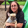 woman, dog, french_bulldog, glasses, playground, slide, harness, leash, outdoor, grass, trees, wood_chips, casual_clothing, smile, pet, holding, sitting, person, animal, park