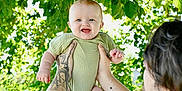 Ava is registered to the contest to win money with this photo: baby, child, tattoos, arms, green_onesie, smiling, outdoor, sunlight, leaves, tree, nature, happy, person, portrait, holding, daylight, skin, head, face, man