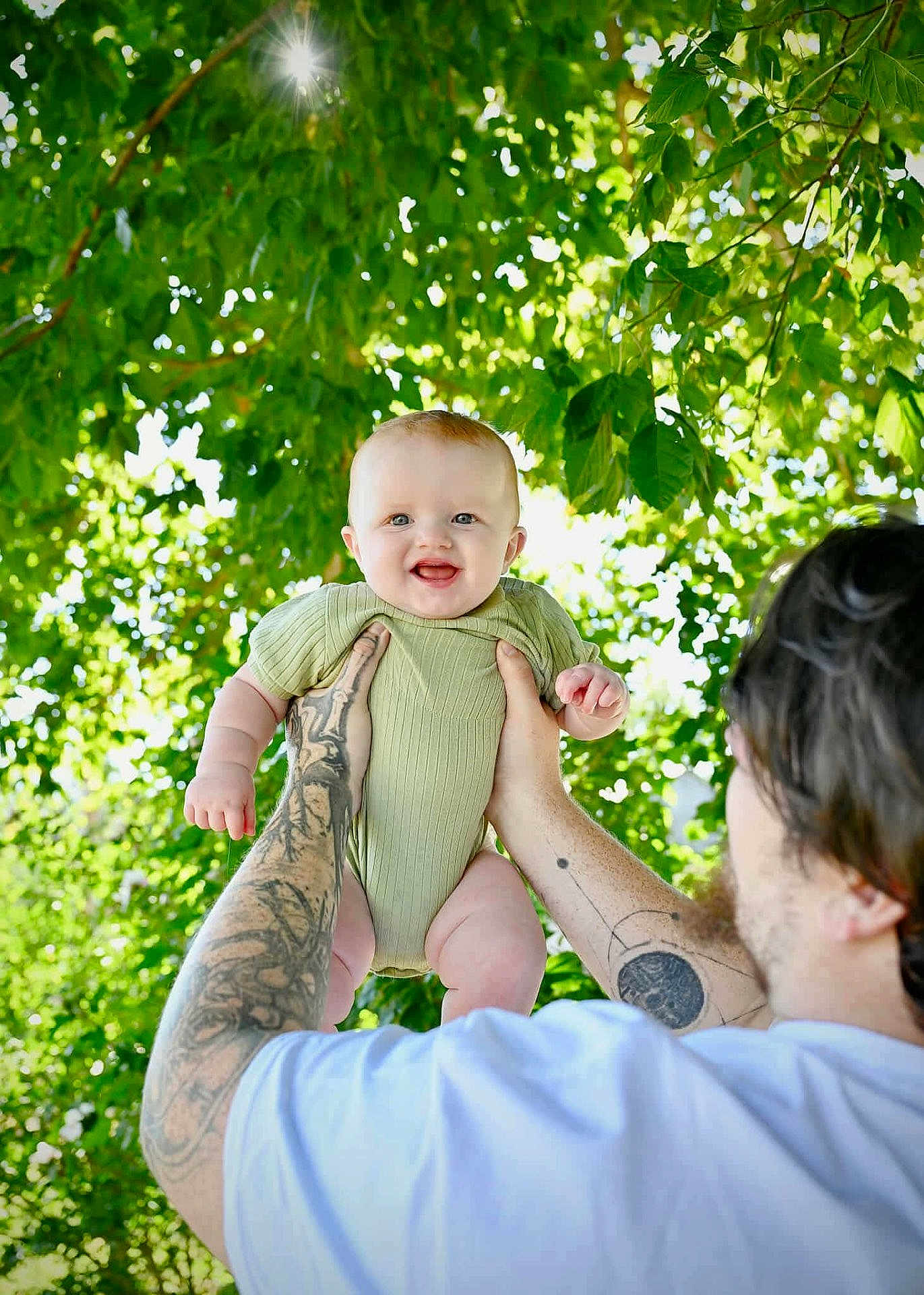 Ava is registered to the contest to win money with this photo: baby, child, tattoos, arms, green_onesie, smiling, outdoor, sunlight, leaves, tree, nature, happy, person, portrait, holding, daylight, skin, head, face, man