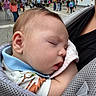 baby, infant, sleeping, stroller, outdoor, street, festival, crowd, portrait, close_up, peaceful, blanket, clothing, parent, carrier, urban, daylight, background, family, child