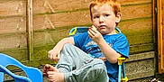 Tobias is registered to the contest to win money with this photo: barefoot, blue_shirt, casual_clothing, child, daylight, garden, grass, gray_pants, outdoor, pensive, plastic_chair, play_area, red_hair, relaxed, seated, small_chair, snack, wooden_deck, wooden_fence, young_boy