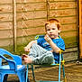 barefoot, blue_shirt, casual_clothing, child, daylight, garden, grass, gray_pants, outdoor, pensive, plastic_chair, play_area, red_hair, relaxed, seated, small_chair, snack, wooden_deck, wooden_fence, young_boy