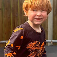 Tobias joined the competition — help win amazing prizes! child, red_hair, dinosaur_shirt, black_shirt, orange_print, smiling, outdoor, fence, wooden_fence, casual_clothing, young_child, happy, portrait, playful, sunlight, daylight, person, kid, long_sleeve_shirt, fun