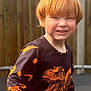 child, red_hair, dinosaur_shirt, black_shirt, orange_print, smiling, outdoor, fence, wooden_fence, casual_clothing, young_child, happy, portrait, playful, sunlight, daylight, person, kid, long_sleeve_shirt, fun