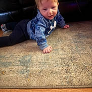 Sutton is registered to the contest to win money with this photo: baby, child, crawling, blue_hoodie, carpet, floor, leather_couch, indoor, wooden_floor, curly_hair, casual_clothing, person, young_child, smiling, hand, face, head, socks, cozy, home