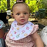 baby, child, infant, pink_bib, pink_dress, bow, curly_hair, outdoor, greenery, bicycle, person, face, cute, young_child, summer, daylight, portrait, sitting, attention, expression