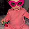 baby, pink_onesie, curly_hair, pacifier, toy, glasses, indoor, bed, cute, child, novelty_glasses, sitting, infant, face, person, playful, cozy, soft_lighting, expression, young_child