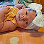 baby, infant, face, headband, bow, yellow_clothing, blanket, orange, heart_pattern, hand, skin, closeup, lying_down, indoors, cute, newborn, soft_texture, portrait, child, peaceful