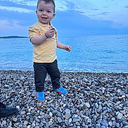 Aris Stefan participe au concours pour gagner de l'argent avec cette photo : beach, blue_shoes, casual_clothing, child, clouds, daytime, happy, holding, nature, ocean, outdoor, pebbles, rock, shore, sky, smiling, standing, toddler, water, yellow_shirt