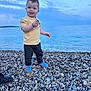 beach, blue_shoes, casual_clothing, child, clouds, daytime, happy, holding, nature, ocean, outdoor, pebbles, rock, shore, sky, smiling, standing, toddler, water, yellow_shirt
