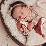 baby, sleeping, infant, plush_toy, heart_shaped_basket, wooden_basket, lace_clothing, soft_blanket, cozy, cute, child, newborn, headband, hand, face, indoors, peaceful, warm_colors, portrait, resting