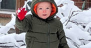 Finn joined the competition — help win amazing prizes! toddler, child, snow, winter_clothing, green_jacket, red_mittens, hood, snowy_background, brick_wall, garden_hose, outdoor, waving, cute, cold_weather, young_child, snow_covered, branches, smiling, person, daylight