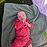 baby, infant, blanket, grass, outdoor, toy, teething_ring, clothing, red_outfit, green_slippers, person, child, cute, lying_down, sunlight, playtime, soft_texture, casual, relaxed, portrait