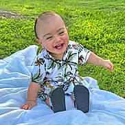 Moses is registered to the contest to win money with this photo: baby, child, smiling, laughing, outdoor, grass, blanket, shirt, tropical_print, footwear, happy, cute, sitting, sunlight, greenery, nature, portrait, infant, casual_clothing, daytime