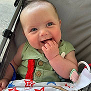 Jett is registered to the contest to win money with this photo: baby, child, smiling, stroller, blue_eyes, hand, clothing, fabric, pattern, red_stars, blue_stars, baseball, toy, green_shirt, happy, cute, infant, portrait, closeup, innocence