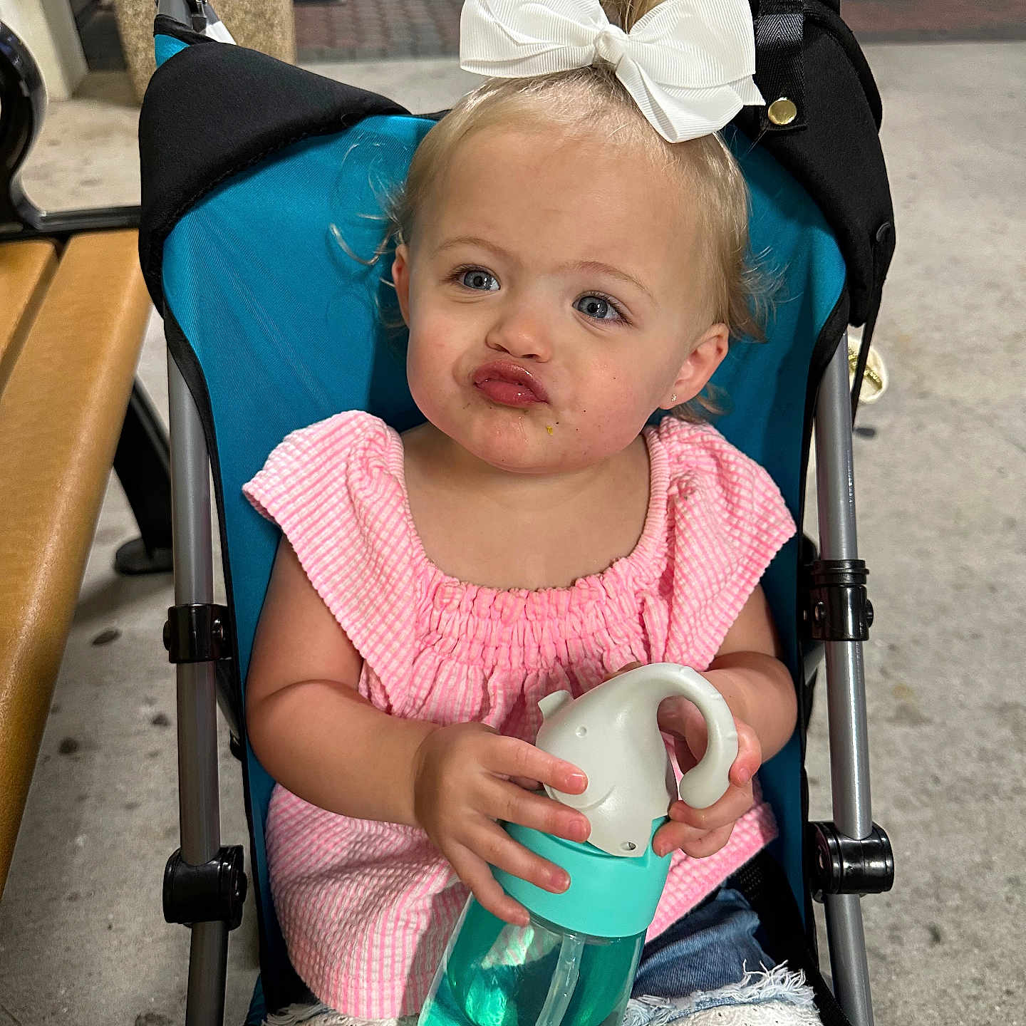 Raegan is registered to the contest to win money with this photo: baby, bench, bow, child, concrete, cute, denim_shorts, expression, holding, lace_trim, outdoor, person, pink_top, playful, pouting_face, sitting, stroller, summer_clothing, toddler, water_bottle