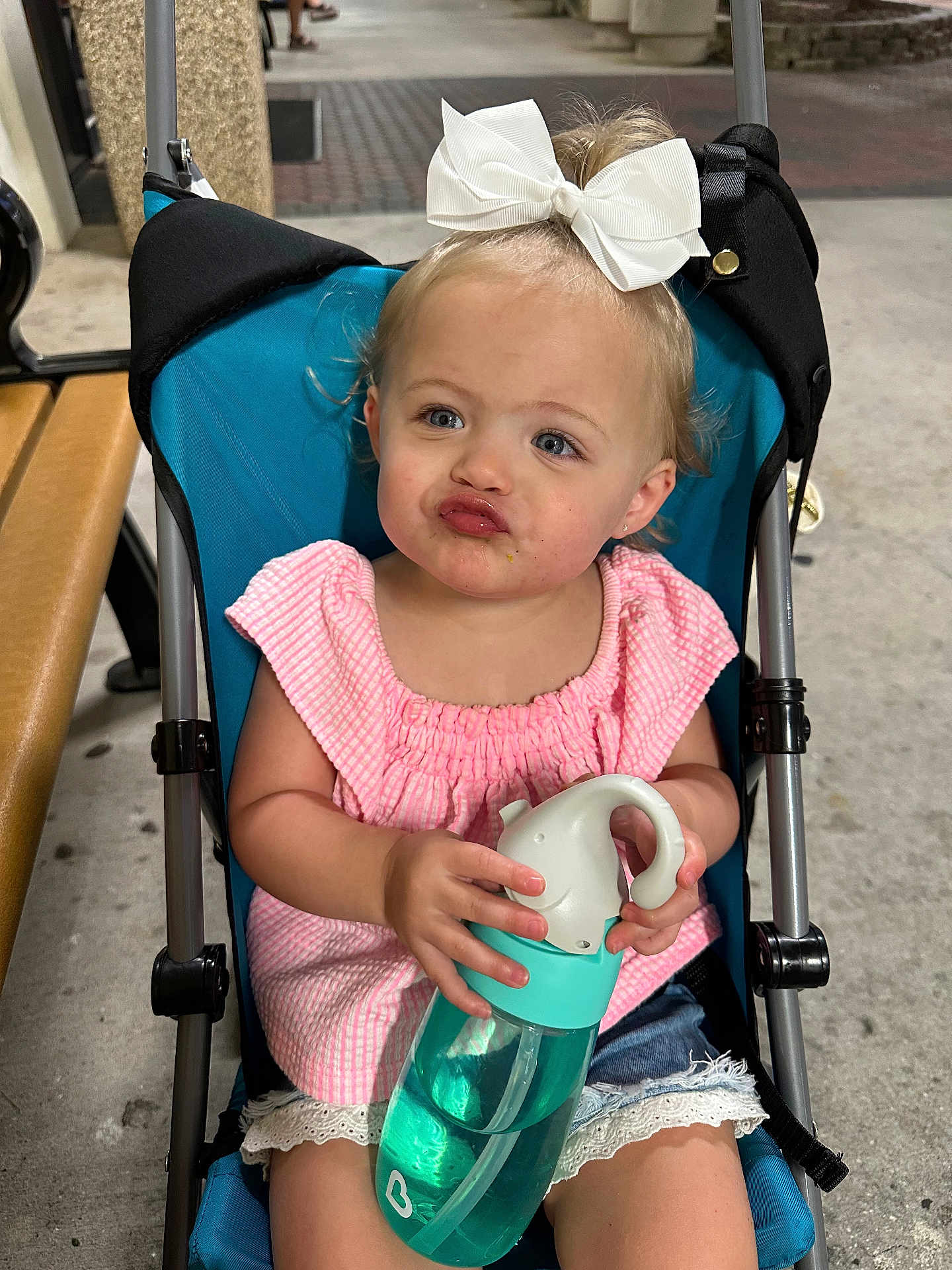 Raegan is registered to the contest to win money with this photo: toddler, child, stroller, bow, water_bottle, pink_top, denim_shorts, lace_trim, pouting_face, outdoor, bench, concrete, cute, baby, person, summer_clothing, expression, sitting, holding, playful