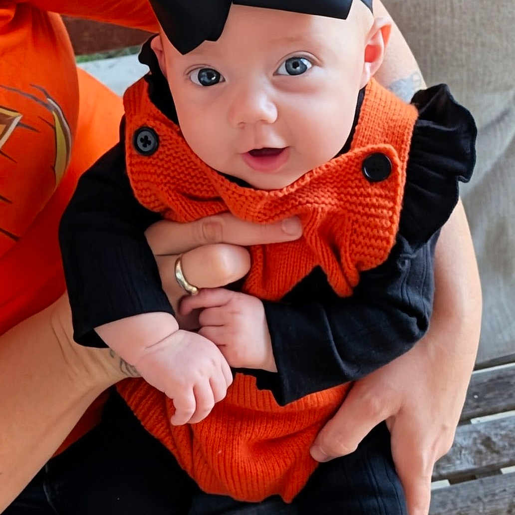 Ryleigh is registered to the contest to win money with this photo: adult, baby, black_bow, black_pants, child, clothing, cute, face, hands, happy, holding, indoor, infant, knitwear, orange_outfit, person, portrait, pumpkin_socks, seated, smiling