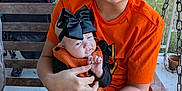 Ryleigh is registered to the contest to win money with this photo: child, baby, smiling, orange_shirt, black_bow, outdoor, bench, stroller, baby_clothing, holding, happy, portrait, greenery, fence, daylight, person, casual_clothing, family, sibling, cute