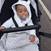 Saint Given joined the competition — help win amazing prizes! baby, black_stroller, blanket, casual, child, comfort, cozy, curious, cute, expression, hoodie, infant, outdoor, pacifier, pavement, small_hand, soft_texture, stroller, white_clothing, young_child