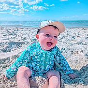 Colton is registered to the contest to win money with this photo: baby, child, beach, sand, ocean, sky, blue_sky, cap, smiling, happy, sunny, vacation, summer, sitting, swimsuit, floral_pattern, outdoor, water, nature, portrait