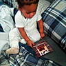 Fasha Douglas is registered to the contest to win money with this photo: blanket, book, card, casual, child, couch, cozy, furniture, hair_braids, home, indoor, pattern, person, plaid, reading, sitting, soft_fabric, toddler, white_clothing, young_child