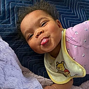 Kalani is registered to the contest to win money with this photo: baby, child, tongue_out, smiling, face, bib, cartoon, blanket, purple_clothing, soft_texture, hand, cute, playful, indoors, infant, closeup, happy, person, head, expression