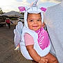 Kalani is registered to the contest to win money with this photo: baby, child, unicorn_costume, costume, wings, smiling, outdoor, daytime, parking_lot, person_holding, car, clouds, sky, hood, pink, white, cute, happy, infant, casual