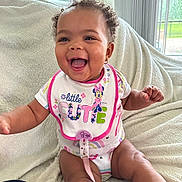 Kalani joined the competition — help win amazing prizes! baby, bib, blanket, bright, child, clothing, colorful, couch, curly_hair, cute, earrings, happy, home, indoors, natural_light, person, sitting, smiling, toddler, window
