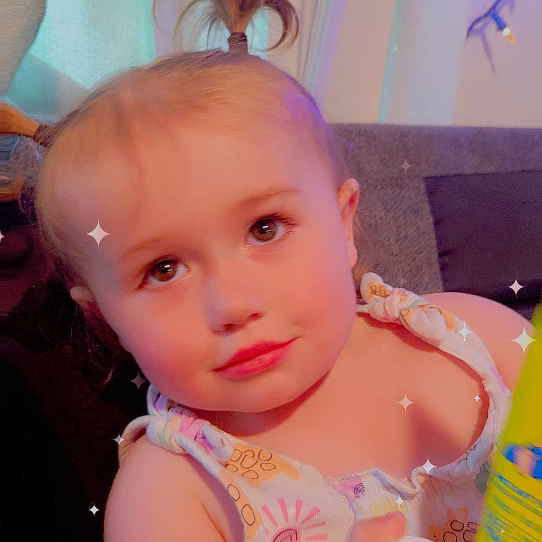 Atalli is registered to the contest to win money with this photo: bottle, child, clothing, couch, curtain, expression, face, hair, hand, indoors, light, living_room, person, pillow, pony_tail, skin, sleeveless, sparkles, toddler, window