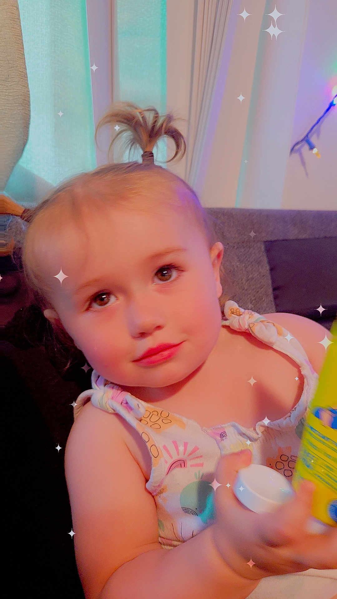 Atalli is registered to the contest to win money with this photo: toddler, child, face, hair, pony_tail, clothing, sleeveless, bottle, hand, living_room, couch, pillow, window, curtain, light, sparkles, skin, expression, indoors, person