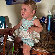 Leia is registered to the contest to win money with this photo: toddler, child, table, hand, romper, shoe, floor, chair, poster, water_bottle, kitchen, person, smile, blue_eyes, earring, indoor, holding_hands, marble_table, tile_floor, clothing