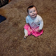 Jazmine is registered to the contest to win money with this photo: baby, child, carpet, indoor, pink_skirt, white_cardigan, sitting, smiling, face, person, floor, shelf, books, clothing, happy, cute, young, portrait, home, carpeted_floor