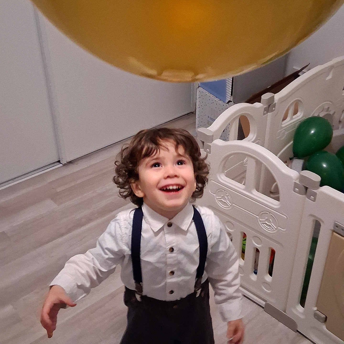 Valentim a rejoint le concours — aidez-le/la à gagner de superbes lots ! balloon, celebration, child, clothing, curly_hair, golden_balloon, green_balloons, happy, home, indoor, joyful, looking_up, person, playpen, smiling, suspenders, toddler, white_shirt, wooden_floor, young_child