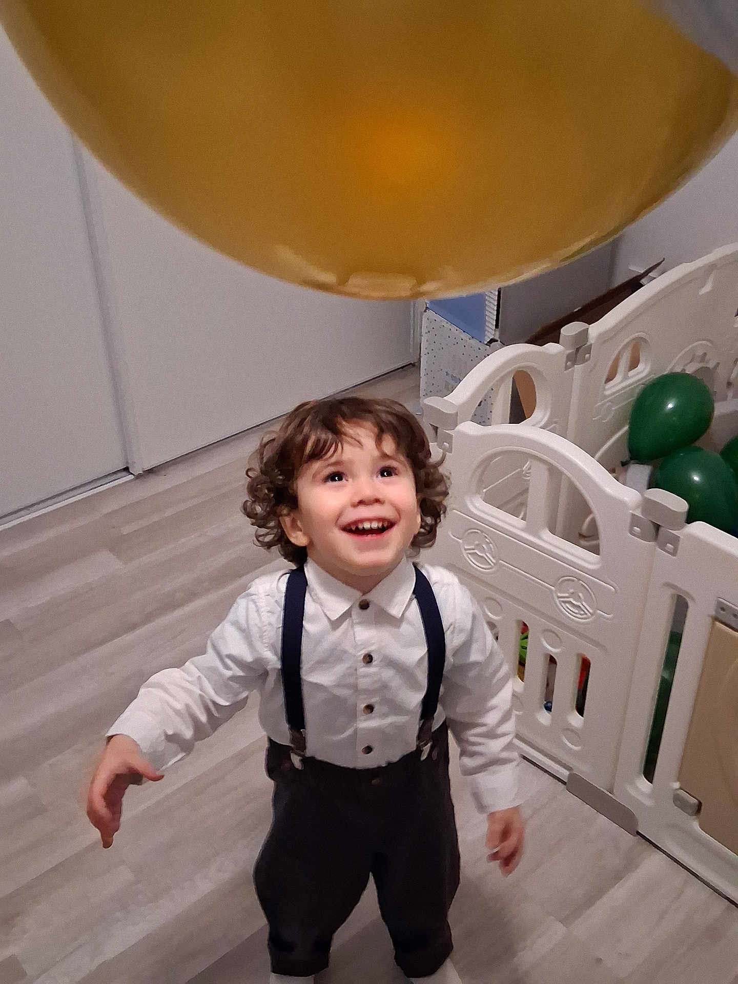Valentim a rejoint le concours — aidez-le/la à gagner de superbes lots ! toddler, child, curly_hair, suspenders, white_shirt, balloon, golden_balloon, indoor, playpen, green_balloons, wooden_floor, happy, smiling, looking_up, young_child, person, clothing, home, celebration, joyful