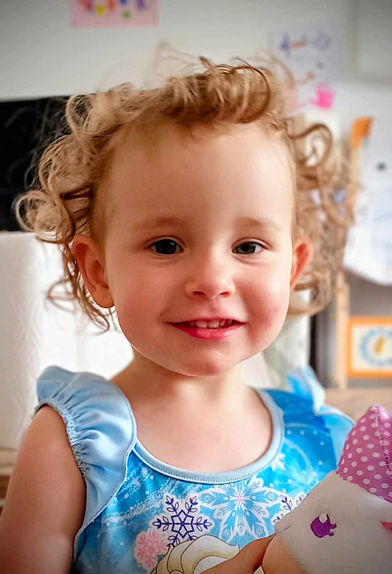 Aïna participe au concours pour gagner de l'argent avec cette photo : child, toddler, smiling, curly_hair, blue_dress, snowflake_pattern, plush_toy, indoor, portrait, cute, happy, young_child, face, person, holding, soft_toy, colorful, casual_clothing, closeup, bright