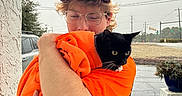Boo Boo is registered to the contest to win money with this photo: person, cat, black_cat, glasses, cap, orange_hoodie, hugging, outdoor, potted_plant, wall, car, road, overcast, sidewalk, animal, pet, affection, young_adult, casual_clothing, nature