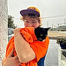person, cat, black_cat, glasses, cap, orange_hoodie, hugging, outdoor, potted_plant, wall, car, road, overcast, sidewalk, animal, pet, affection, young_adult, casual_clothing, nature