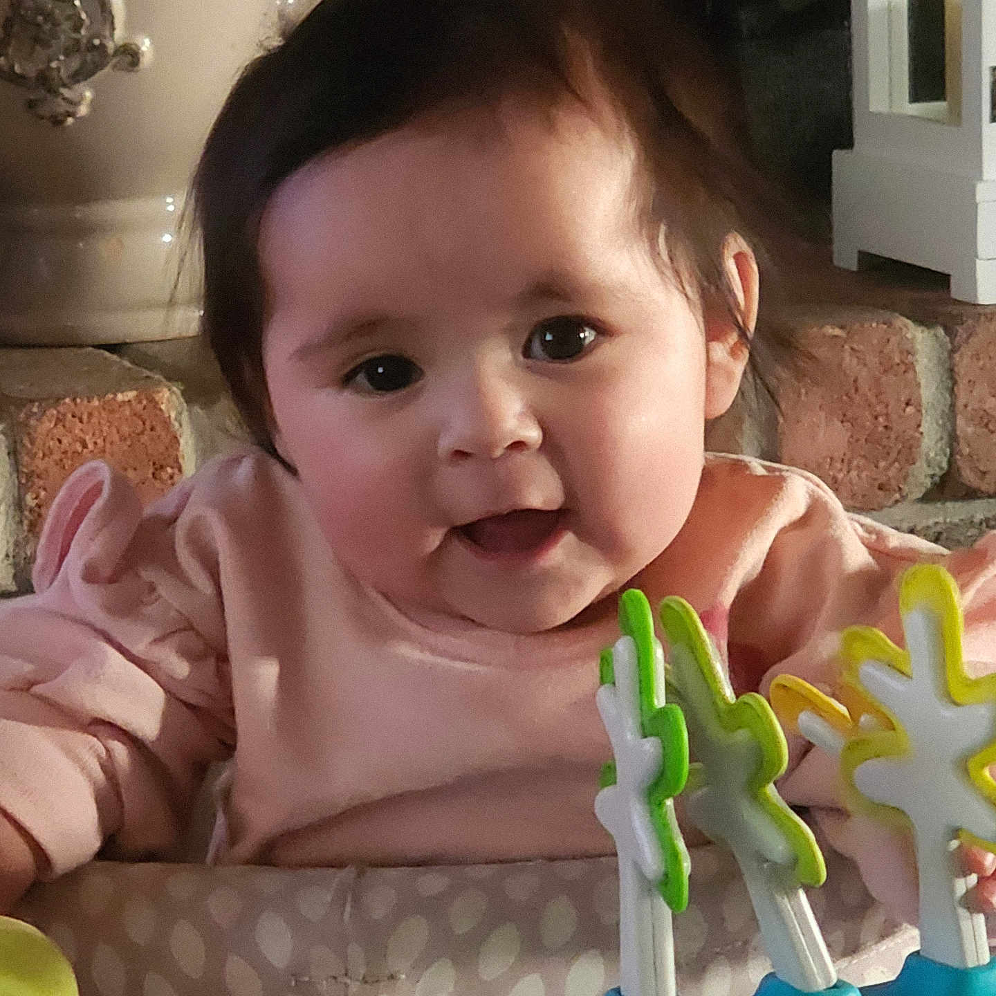 Sofia is registered to the contest to win money with this photo: baby, child, infant, face, smile, pink_clothing, toy, indoor, home_decor, brick_wall, curly_hair, cute, playful, young_child, cheerful, person, portrait, closeup, bright_eyes, cozy
