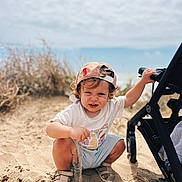 Roméo participe au concours pour gagner de l'argent avec cette photo : beach, cap, child, curly_hair, daylight, footwear, grass, nature, outdoor, person, playing, sand, shorts, sky, smiling, squatting, stroller, sunny, toddler, tshirt